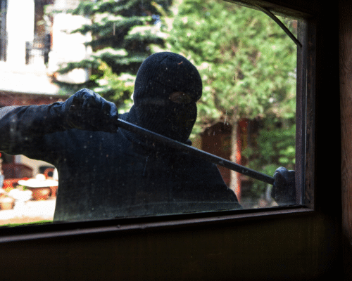 Masked individual wearing black gloves attempting to pry open a window with a crowbar, symbolizing San Diego burglary defense cases and the legal differences between residential and commercial break-ins under California Penal Code 459 and 460.