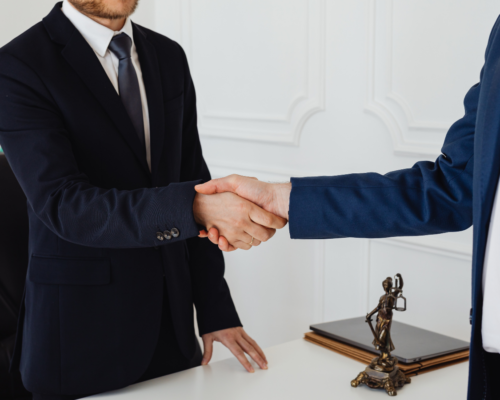 Two individuals in business attire shake hands across a desk, symbolizing legal representation and partnership, with a small Lady Justice statue and documents visible in a professional office setting.