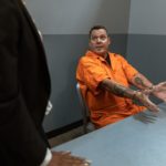 Man in a prison jumpsuit in an interrogation room. He is handcuffed to the table and making an expression. We see the back of a police officer who is facing the criminal.