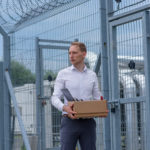 A businessman released from prison holding a box of belongings outside the prison gates.